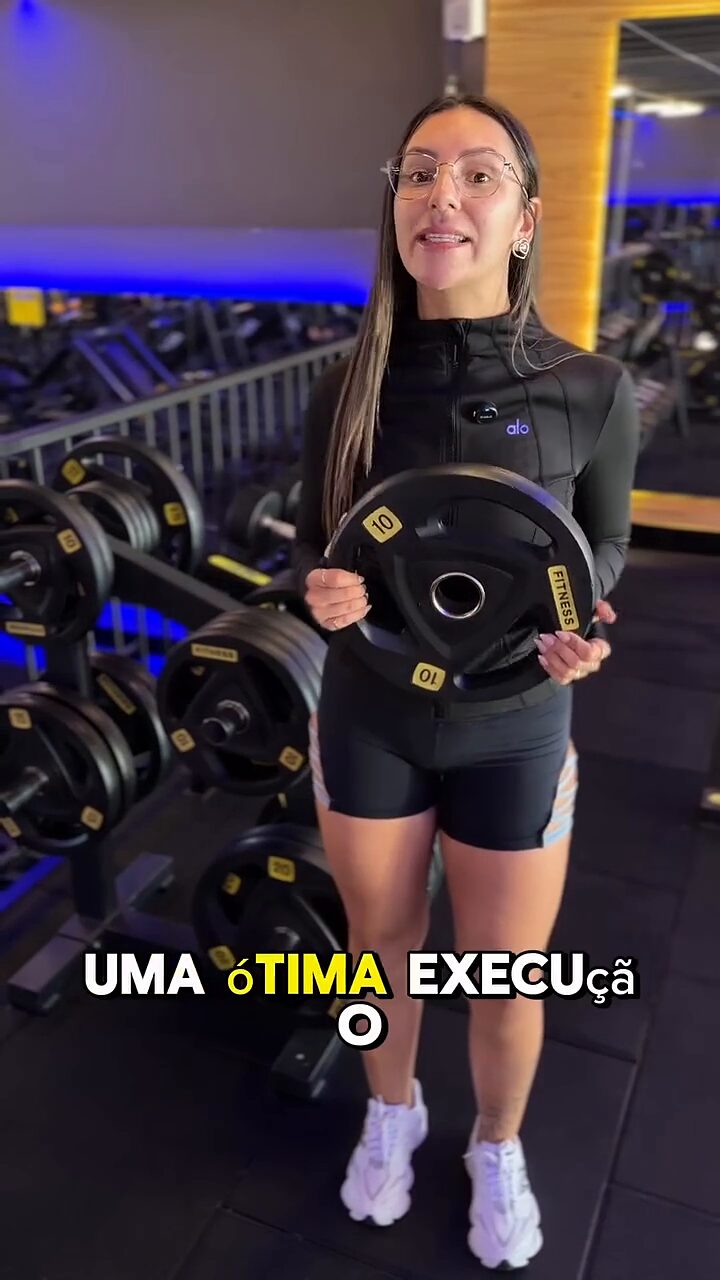 Vitória na academia, segurando uma anilha ao lado do rack de pesos.