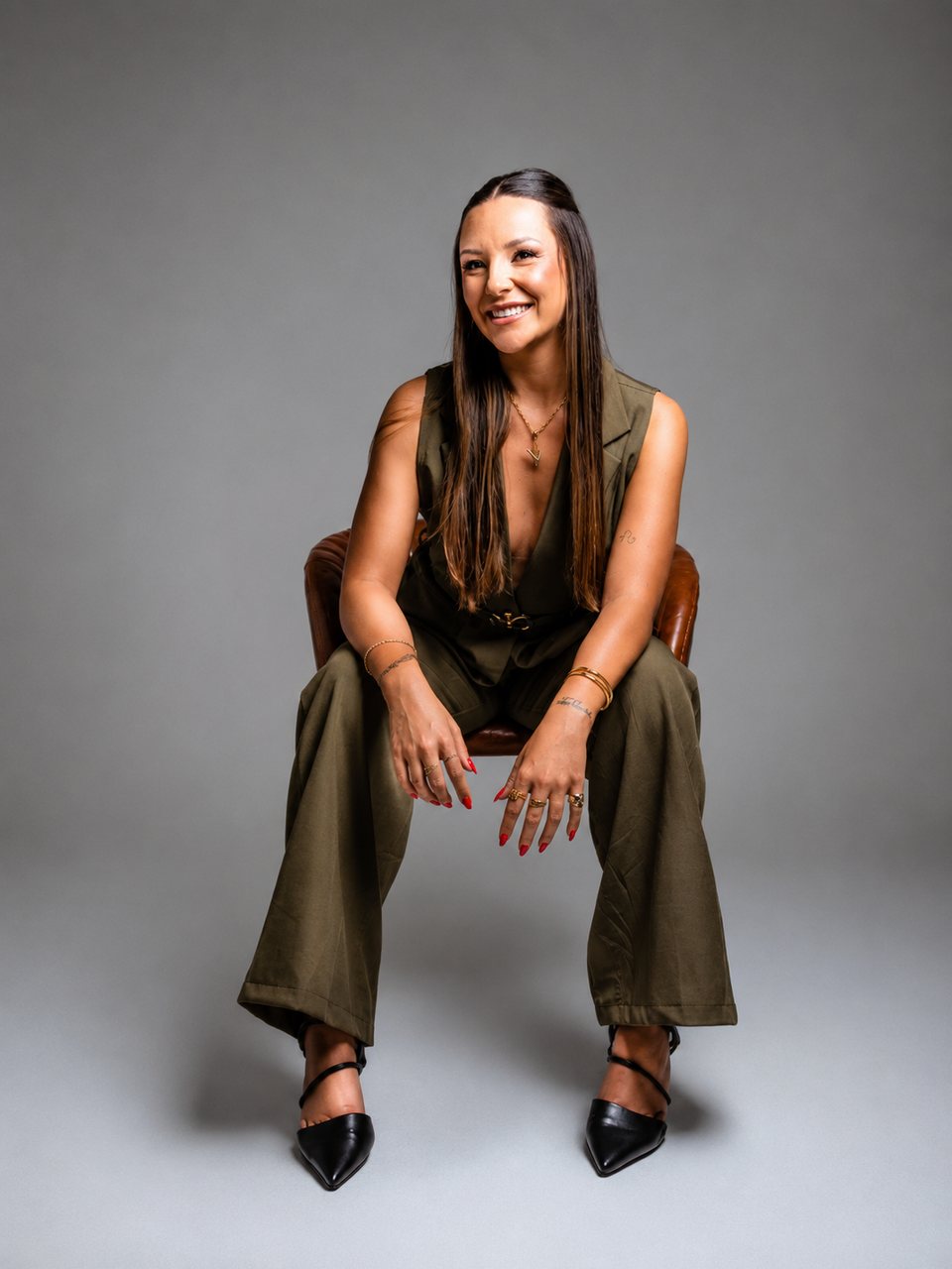 Vitória sentada em uma poltrona de couro, rindo, de conjunto de alfaiataria verde militar.