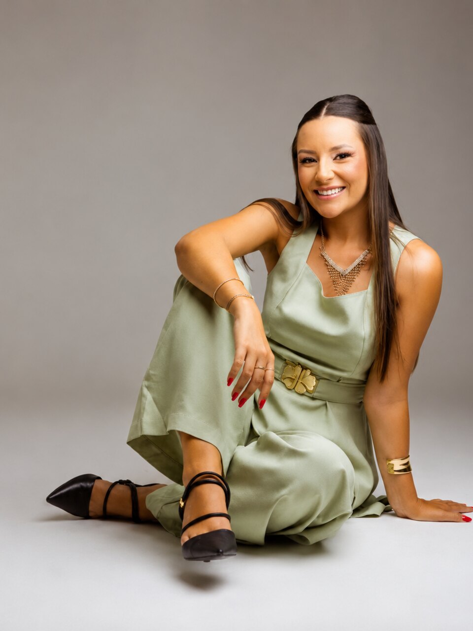 Vitória Claudino sentada no chão do estúdio, sorrindo para a câmera, de vestido verde-claro com cinto dourado.