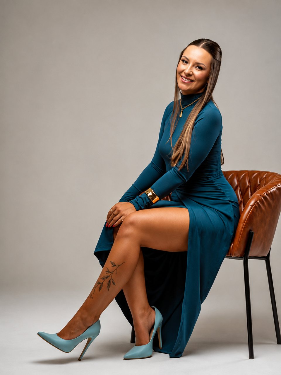 Vitória sentada em uma poltrona caramelo, de vestido azul petróleo de manga longa.
